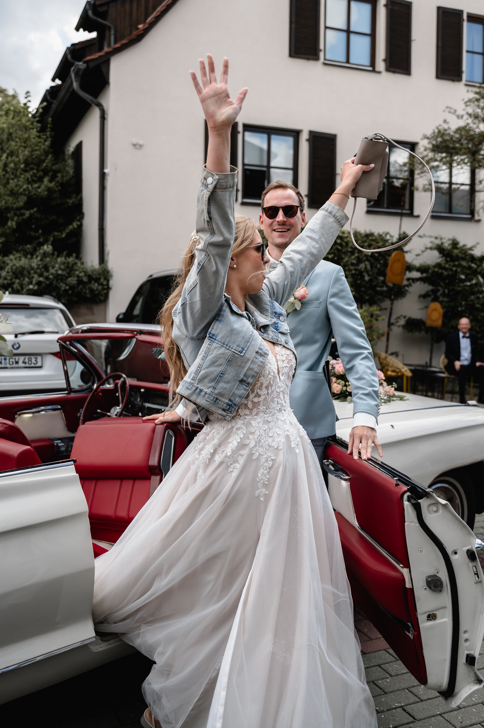 Braut jubelt ihren Hochzeitsgästen am Schindlerhof Nürnberg zu, nachdem sie aus dem Hochzeitsauto ausgestiegen ist. Hochzeitsfoto aufgenommen von der Hochzeitsfotografin Kristina Reinartz